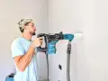 Giovane con capelli biondi usa un trapano Makita per fare un foro nel muro. Il trapano non buca muro, ma aspira la polvere.