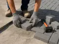 Mani guantate posano con cura le piastrelle grigie, creando un nuovo percorso. Cemento e martello sono pronti per completare il lavoro.