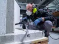 Uomo con mascherina, occhiali e cuffie protettive usa un martello perforatore per forare cemento armato.