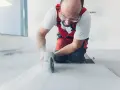 Operaio con occhiali protettivi e tuta rossa mentre impara come tagliare gres porcellanato con una smerigliatrice angolare, sollevando polvere.