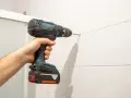 Mano che usa un trapano per forare le piastrelle bianche. Un'ombra lunga si proietta sul muro.
