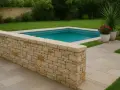 Un muretto in pietra fai da te delimita una piscina azzurra, circondata da prato verde e pavimentazione chiara.