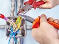 Mani esperte usano **le tenaglie** per lavorare sui fili elettrici in una scatola di derivazione, con tubi corrugati sullo sfondo.