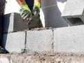 Mani guantate stendono cemento per coprire un muro esterno di cemento, posando blocchi.