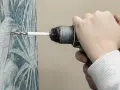 Mano che usa un trapano per forare un muro con carta da parati decorata, evitando di rovinare l'intonaco.