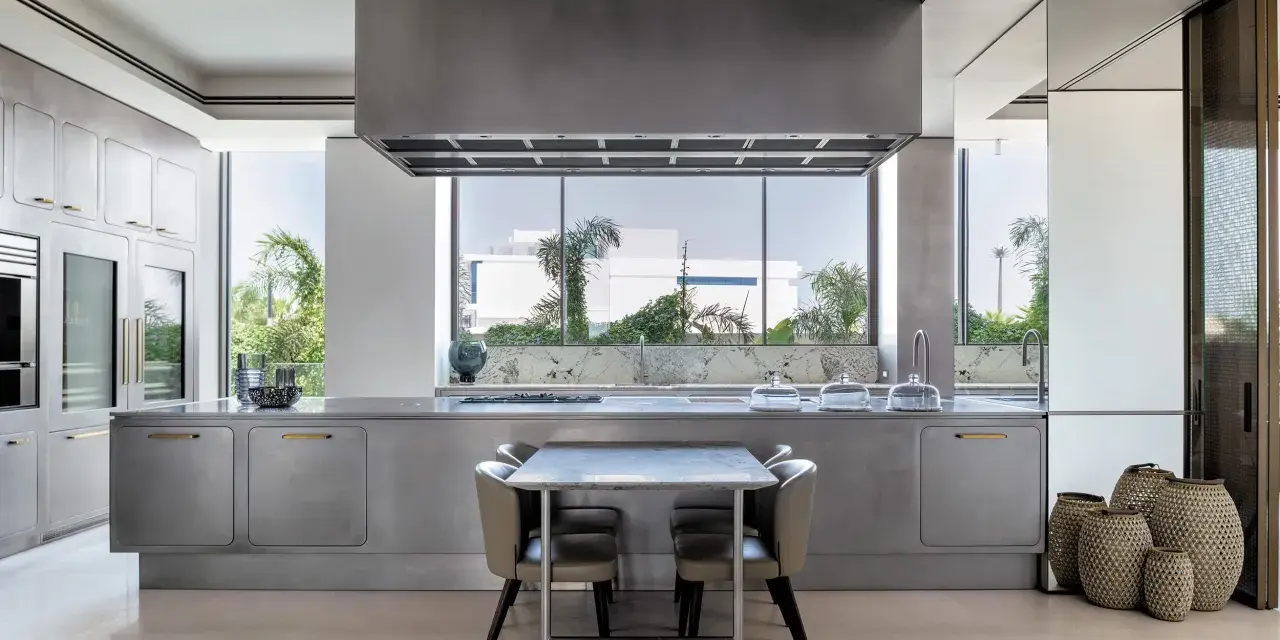 Cucina moderna con isola centrale in acciaio opaco, piano in marmo, tavolo e sedie. Ampia finestra con vista sul verde.