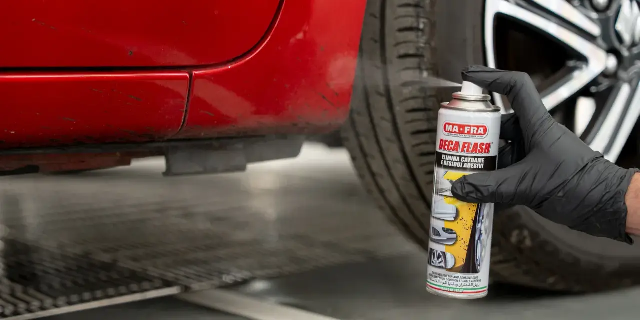 Mano con guanto nero spruzza spray "Deca Flash" su un'auto rossa per come sciogliere la colla secca e residui adesivi.
