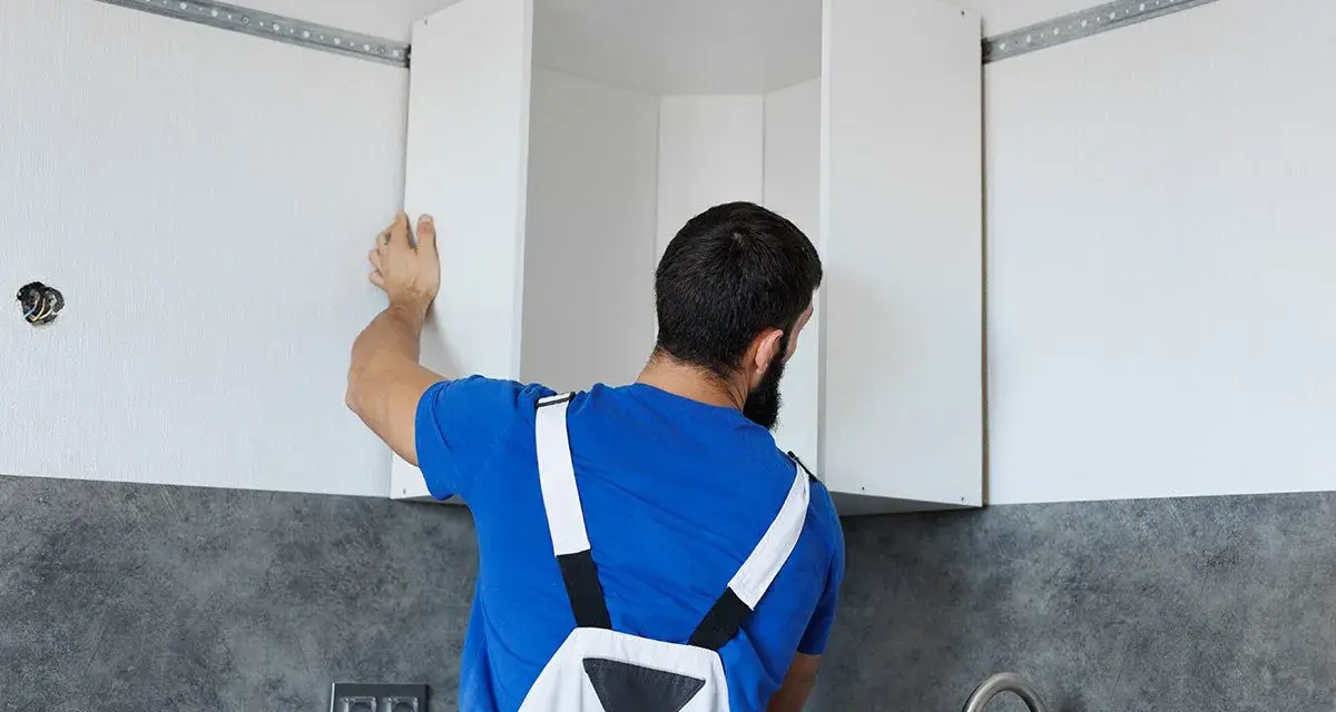 Installazione di pensili da cucina: un uomo con tuta blu monta un pensile angolare su una guida a muro.