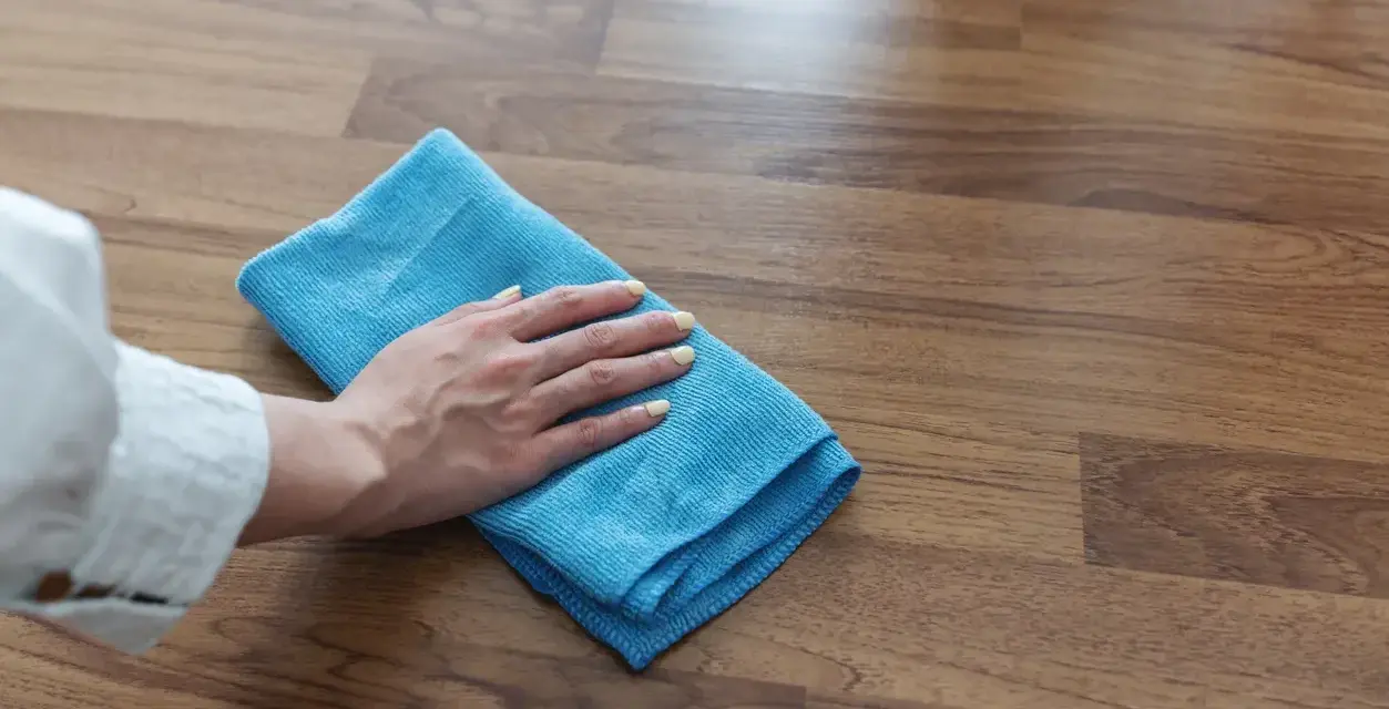 Mano con panno blu pulisce superficie in legno, mostrando come togliere la colla dal legno con delicatezza.