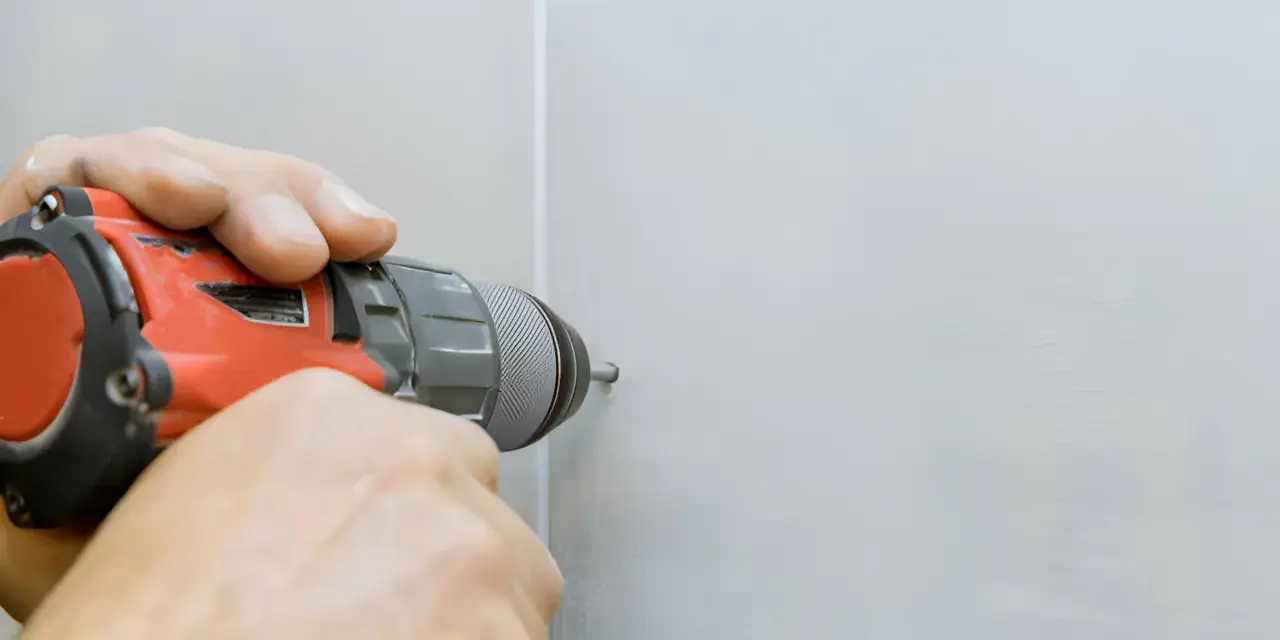 Mani che usano un trapano per fare un buco nel muro bianco.