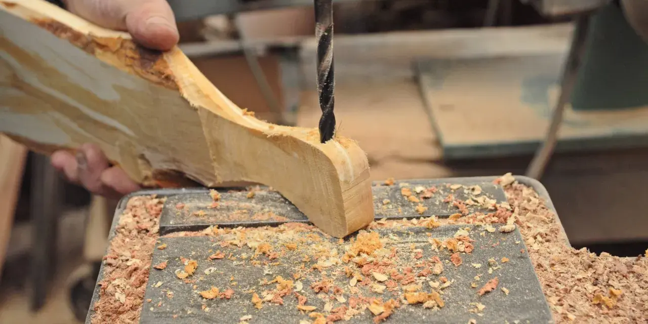 Mano che trapana un pezzo di legno grezzo, creando un tagliere legno fai da te. Segatura ovunque.