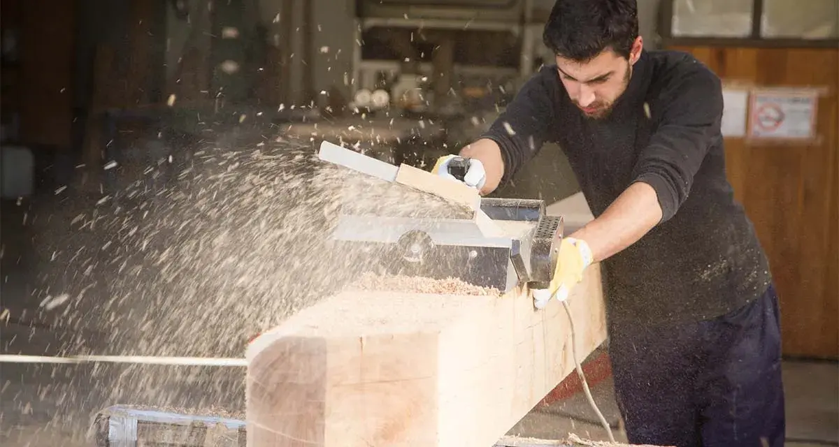 Falegname usa pialla elettrica su un grosso tronco di legno, creando una nuvola di segatura. Il piallare significato è dare una finitura liscia.