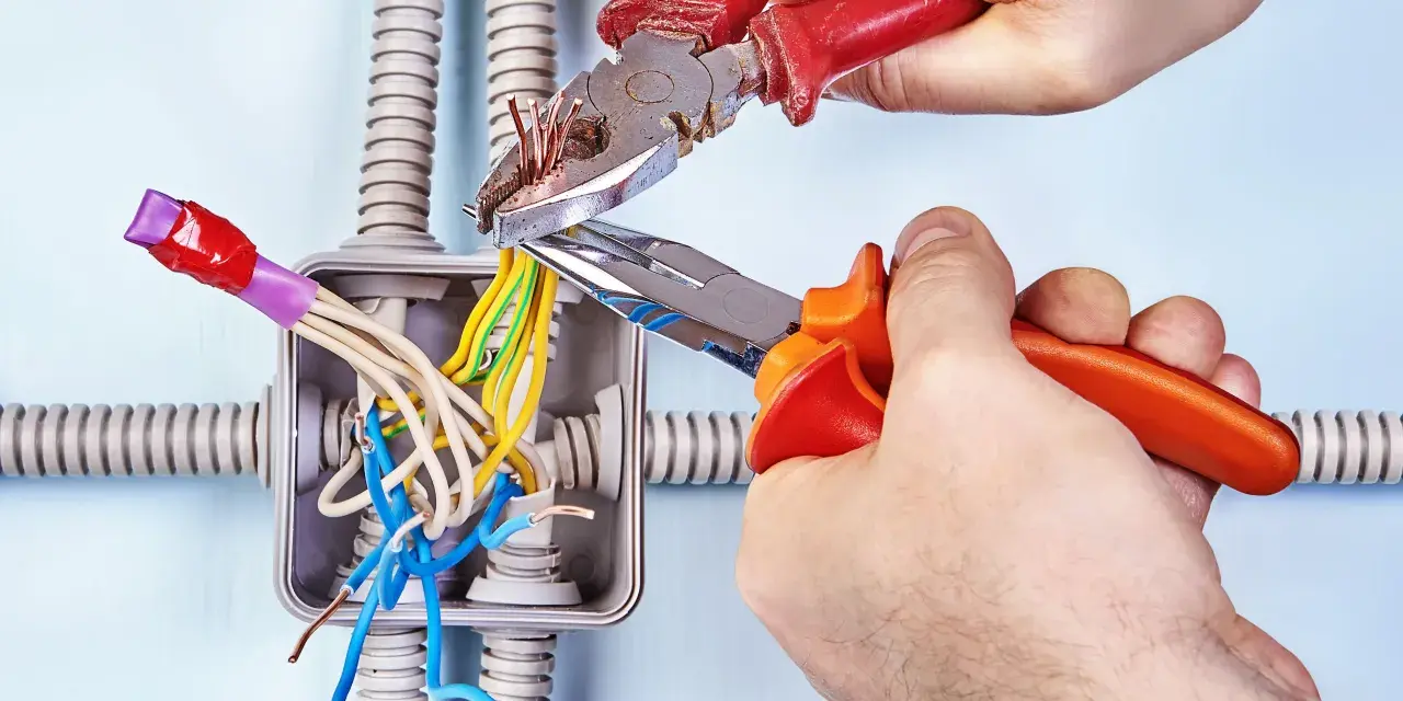 Mani esperte usano **le tenaglie** per lavorare sui fili elettrici in una scatola di derivazione, con tubi corrugati sullo sfondo.