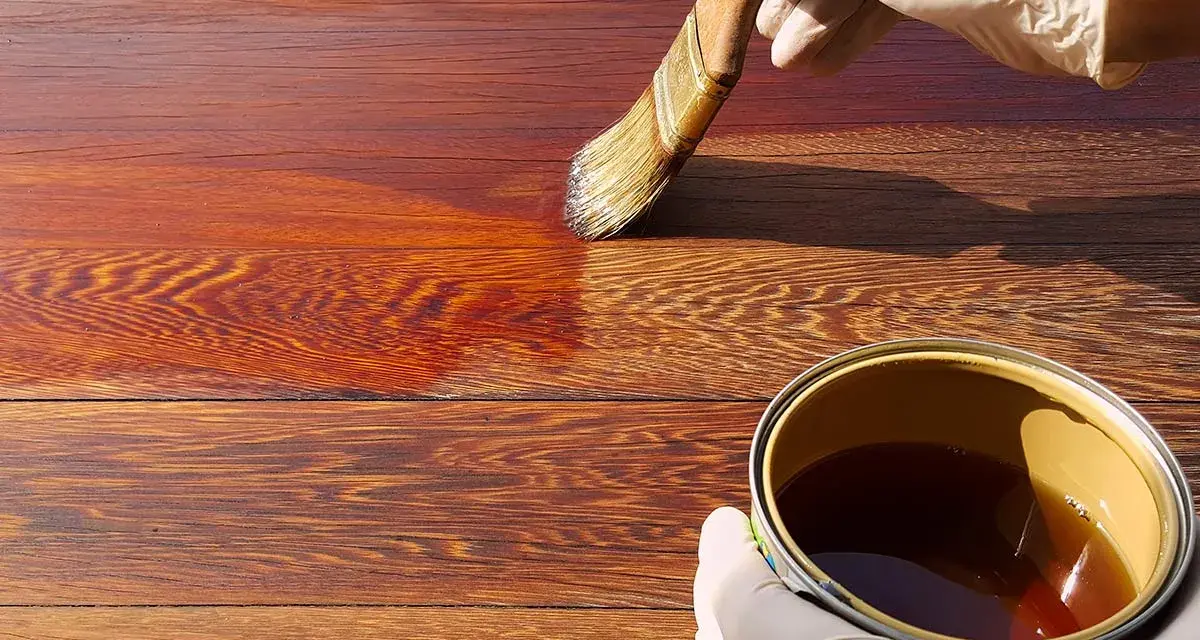 Mano guantata applica una vernice scura su assi di legno, mostrando come scurire il legno per un aspetto più profondo e ricco.