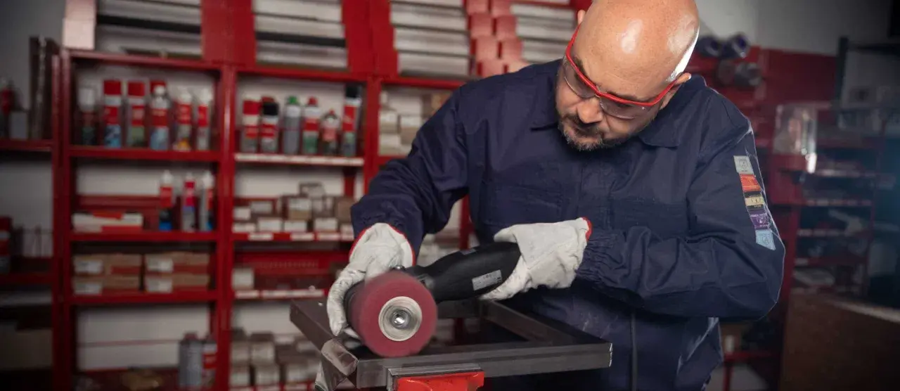 Operaio con occhiali protettivi e guanti lavora su un pezzo metallico, usando una smerigliatrice. Impara come saldare acciaio inox con precisione.