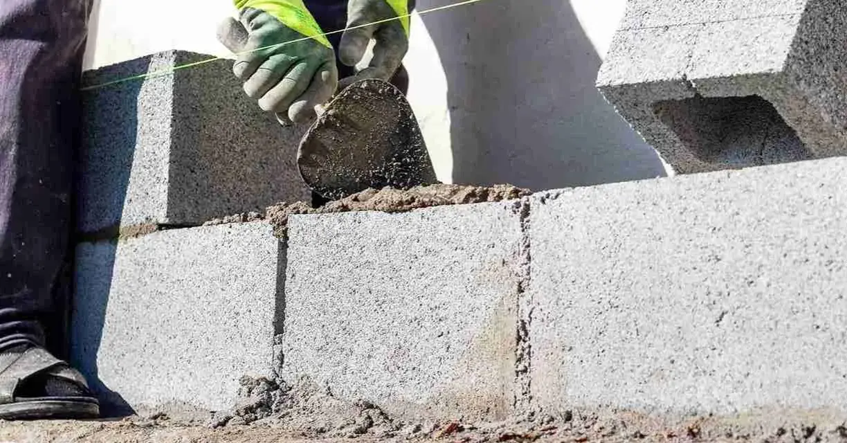 Mani guantate stendono cemento per coprire un muro esterno di cemento, posando blocchi.