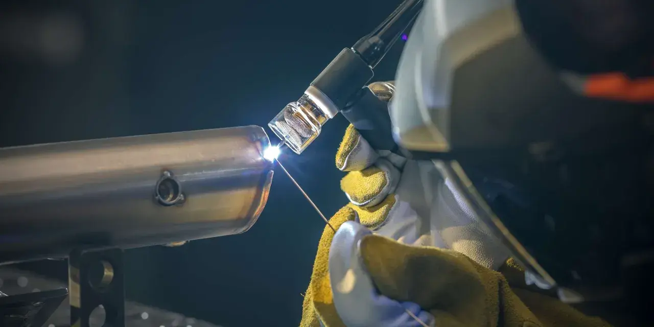 Saldatura TIG di un tubo metallico, con fiamma brillante e filo di apporto. L'artigiano indossa guanti protettivi gialli.