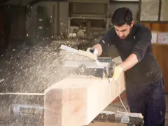 Falegname usa pialla elettrica su un grosso tronco di legno, creando una nuvola di segatura. Il piallare significato è dare una finitura liscia.