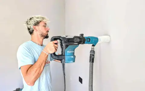 Giovane con capelli biondi usa un trapano Makita per fare un foro nel muro. Il trapano non buca muro, ma aspira la polvere.