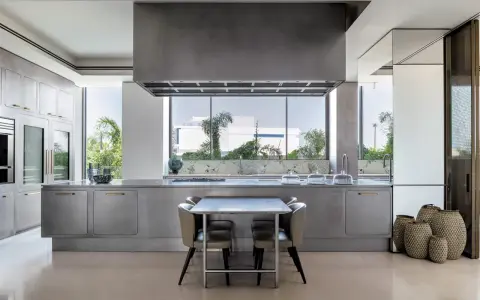 Cucina moderna con isola centrale in acciaio opaco, piano in marmo, tavolo e sedie. Ampia finestra con vista sul verde.