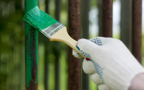 Mano guantata che vernicia di verde un palo di metallo arrugginito, rinnovando la verniciatura.