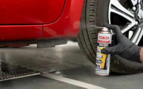 Mano con guanto nero spruzza spray "Deca Flash" su un'auto rossa per come sciogliere la colla secca e residui adesivi.