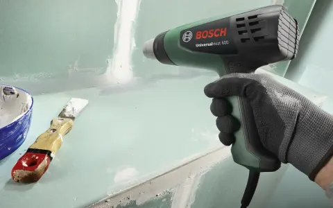 Mano guantata impugna pistola termica Bosch per lavori di finitura. Spatola e stucco a lato.