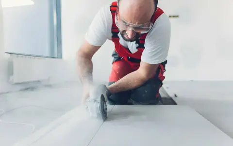 Operaio con occhiali protettivi e tuta rossa mentre impara come tagliare gres porcellanato con una smerigliatrice angolare, sollevando polvere.
