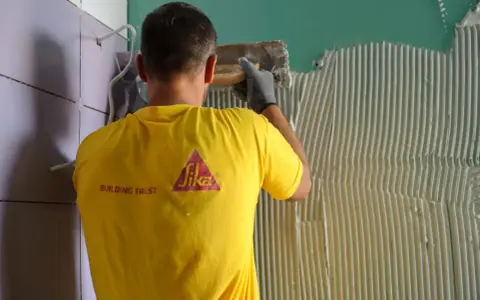 Un operaio con maglia gialla applica colla sul muro, mostrando come attaccare piastrelle.