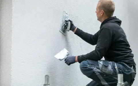 Operaio con cazzuola lavora su un muro esterno, mostrando come rasare un muro rovinato per un aspetto liscio e uniforme.