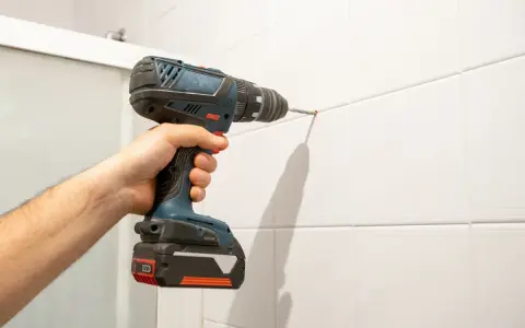 Mano che usa un trapano per forare le piastrelle bianche. Un'ombra lunga si proietta sul muro.