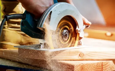 Mano che usa una sega circolare, uno dei tanti tipi di seghe, per tagliare legno. La polvere di legno vola nell'aria.