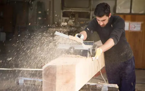 Falegname usa pialla elettrica su un grosso tronco di legno, creando una nuvola di segatura. Il piallare significato è dare una finitura liscia.
