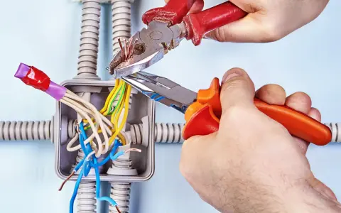 Mani esperte usano **le tenaglie** per lavorare sui fili elettrici in una scatola di derivazione, con tubi corrugati sullo sfondo.