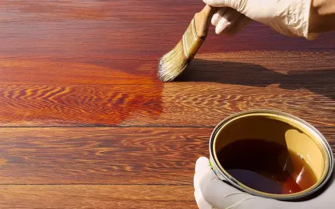 Mano guantata applica una vernice scura su assi di legno, mostrando come scurire il legno per un aspetto più profondo e ricco.