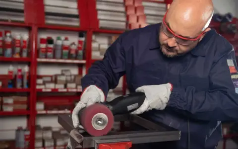 Operaio con occhiali protettivi e guanti lavora su un pezzo metallico, usando una smerigliatrice. Impara come saldare acciaio inox con precisione.