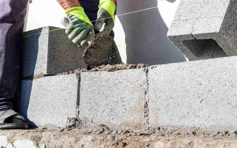 Mani guantate stendono cemento per coprire un muro esterno di cemento, posando blocchi.