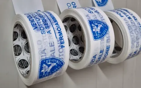 Rotoli di nastri adesivi con scritte blu: "Veicolo Sottosequestro", "Polizia Stradale". Diversi tipi di nastri per imballaggi.
