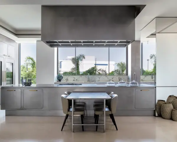 Cucina moderna con isola centrale in acciaio opaco, piano in marmo, tavolo e sedie. Ampia finestra con vista sul verde.