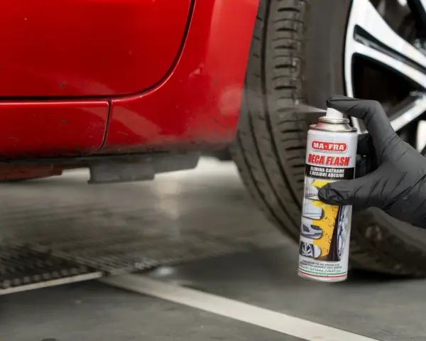 Mano con guanto nero spruzza spray "Deca Flash" su un'auto rossa per come sciogliere la colla secca e residui adesivi.