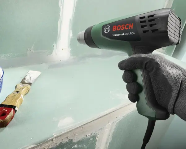 Mano guantata impugna pistola termica Bosch per lavori di finitura. Spatola e stucco a lato.