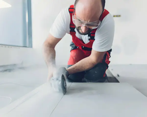 Operaio con occhiali protettivi e tuta rossa mentre impara come tagliare gres porcellanato con una smerigliatrice angolare, sollevando polvere.