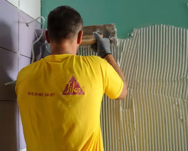 Un operaio con maglia gialla applica colla sul muro, mostrando come attaccare piastrelle.