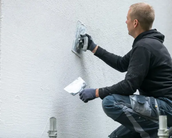 Operaio con cazzuola lavora su un muro esterno, mostrando come rasare un muro rovinato per un aspetto liscio e uniforme.