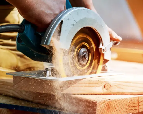 Mano che usa una sega circolare, uno dei tanti tipi di seghe, per tagliare legno. La polvere di legno vola nell'aria.