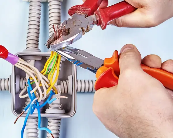 Mani esperte usano **le tenaglie** per lavorare sui fili elettrici in una scatola di derivazione, con tubi corrugati sullo sfondo.