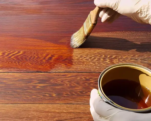 Mano guantata applica una vernice scura su assi di legno, mostrando come scurire il legno per un aspetto più profondo e ricco.