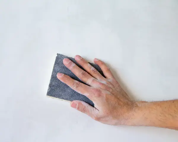 Mano che leviga una parete con carta vetrata, mostrando le sue caratteristiche abrasive per una finitura liscia.