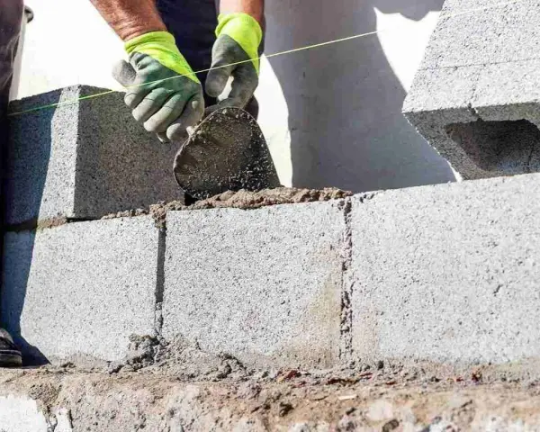 Mani guantate stendono cemento per coprire un muro esterno di cemento, posando blocchi.
