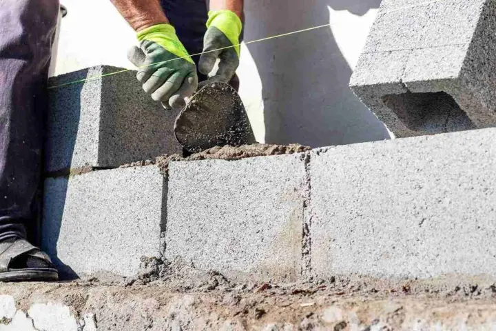 Mani guantate stendono cemento per coprire un muro esterno di cemento, posando blocchi.
